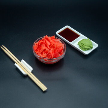 Pickled pink ginger in glass bowl with soy sauce wasabi and chopsticks on dark background top view