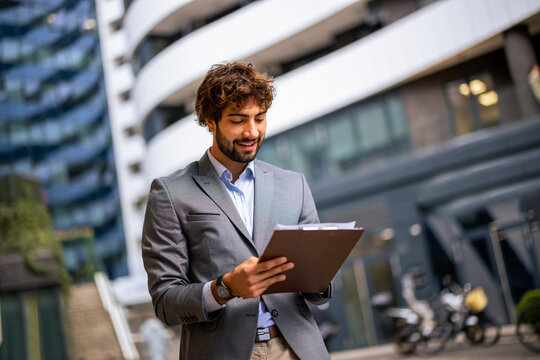 Smiling man in suit holding clipboard outdoors in modern urban office district