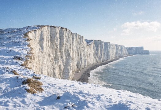 Dramatic winter snowfall covers white chalk cliffs above a stormy sea, with crashing waves, rocky shoreline, dark clouds, and rugged coastal scenery.