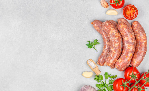 Raw sausages with tomatoes, garlic, onions, parsley, and seasoning arranged on textured gray background with generous empty space for culinary layouts and food photography