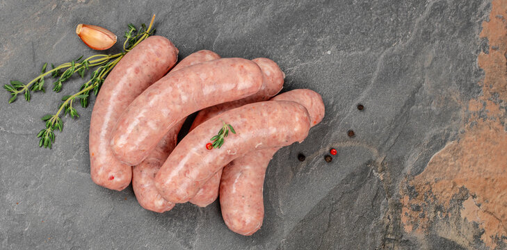 Raw pork sausages arranged with rosemary, peppercorns, and sliced garlic on rustic dark slate surface, overhead food composition showing uncooked meat links