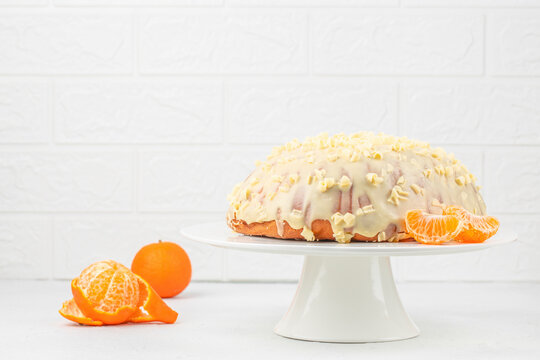 Glazed citrus cake with mandarin segments and whole orange on white pedestal stand against textured brick wall, bright festive dessert still life