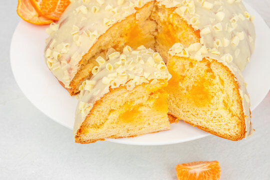Sliced orange cake with white icing and almond flakes arranged on a white plate, accompanied by fresh orange pieces, overhead food still life on a pale surface