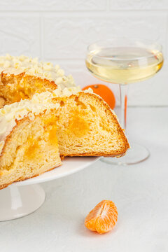 Sliced citrus cake with white chocolate curls beside sparkling drink and peeled orange segment on pedestal plate against textured white wall still life