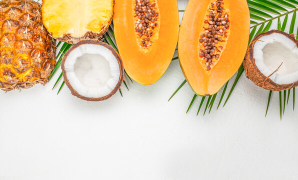 Tropical fruits arranged along upper edge with sliced pineapple, halved papayas, coconut and palm leaves on white background, bright food flat lay with copy space