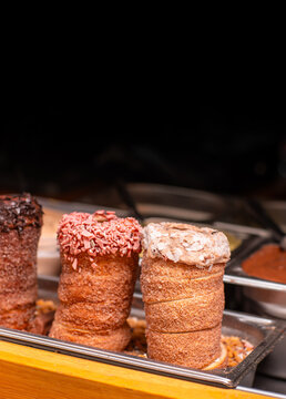 Three cylindrical baked pastries with chocolate, pink and white toppings displayed in metal tray at bakery counter with dark background, closeup food photograph