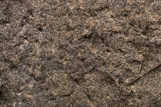 Weathered brown granite stone surface with coarse grain, layered mineral texture, natural cracks, irregular relief, and earthy neutral tones forming a rugged geological background