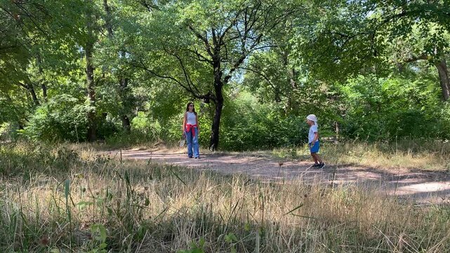 A mother and her young son play badminton on a dirt path in the forest. The active boy holds his racket while his mother watches. They enjoy a cheerful, joyful day outdoors.