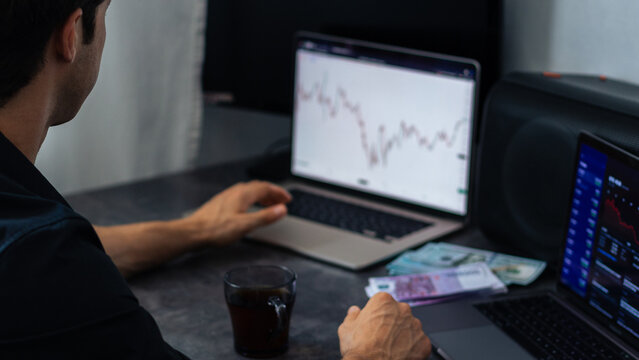 Male trader analyzing financial charts on laptop screens while seated at a desk with coffee and cash notes in a modern workspace