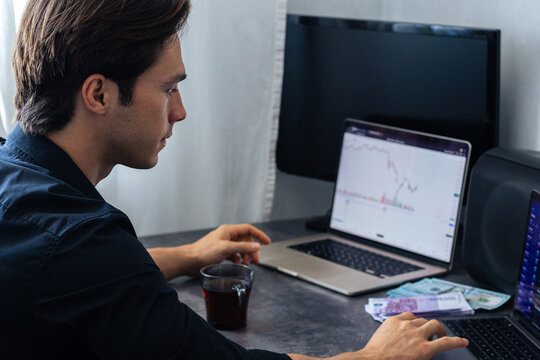 Young male trader analyzing financial charts on laptop while seated at a desk with coffee cup and banknotes in a modern home office setting