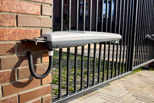 Automatic sliding gate arm on brick pillar with metal chain and black iron gate shadow across tiled driveway in bright daylight entrance security