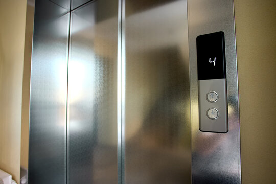 Stainless steel elevator doors closed with illuminated floor indicator panel and brushed metal control buttons in warm interior corridor with soft reflections and vertical seam
