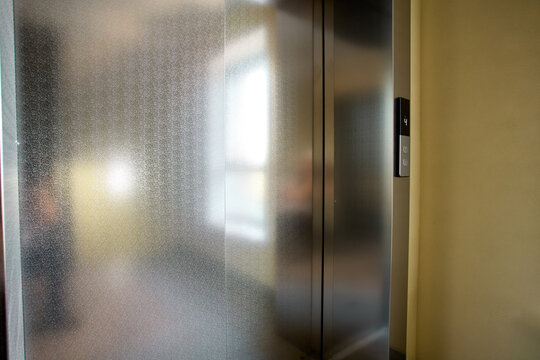 Stainless steel elevator doors closed with illuminated floor indicator panel and brushed metal control buttons in warm interior corridor with soft reflections and vertical seam
