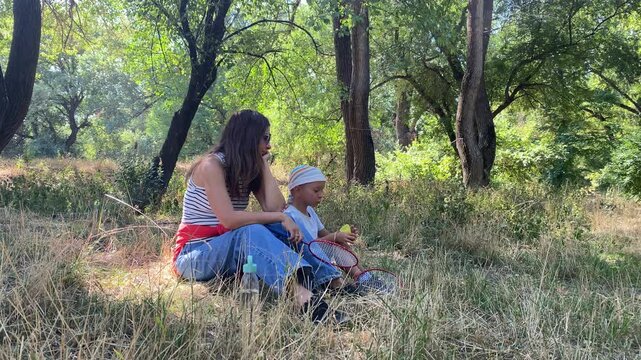 A thoughtful mother and her happy young son sit together on the grass. They rest with their badminton rackets in a sunny green park. The family enjoys a peaceful and contented moment.