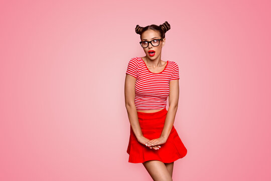 Charming brunette girl with a surprised face coquettishly and shyly holding her hands on a skirt lifting her knee isolated on vivid red background