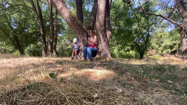 A mother and small child sit happily on folding chairs under big trees, holding toy rackets. They enjoy a peaceful moment together in a sunny park.