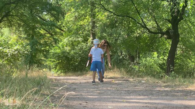 A loving mother bends down to talk with her young son while holding badminton rackets. They share a tender moment on a dirt path surrounded by lush green forest.