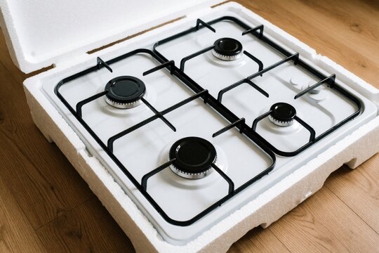 New white gas stove sits in protective foam packaging on wooden floor. Unopened cooktop with black metal grates arrives for kitchen renovation. Fresh appliance delivery ready for installation in