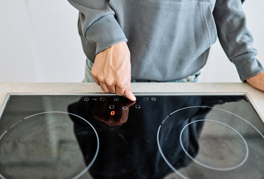 A teenager cautiously testing the heat on a sleek induction cooktop in a modern home kitchen. The image shows a close-up of a hand hovering over the glass-ceramic stove surface, conveying cooking safe