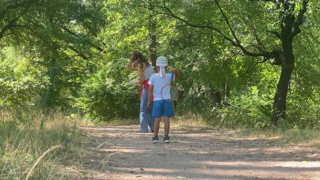 A young child holds a racket high while walking with his loving mother on a dirt path. They enjoy a peaceful game surrounded by green trees. The sunny afternoon brings warmth and cheerful joy.