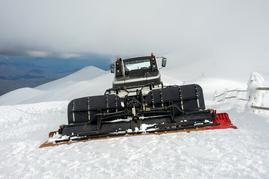 Front view of a heavy duty snowcat groomer machine parked on a frozen alpine mountain slope actively preparing the winter piste terrain for professional skiing and snowboarding.