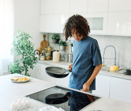 A teen concentrates at a modern kitchen stove, adjusting the cooktop while a frying pan waits on the burner. Casual hoodie, indoor plants and light create a relaxed, everyday domestic scene. Useful fo