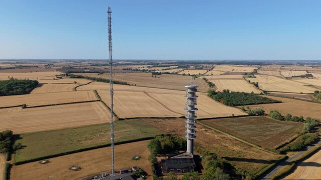 Aerial view of tall telecommunications transmitter mast surrounded by dry summer fields at sunrise in the UK