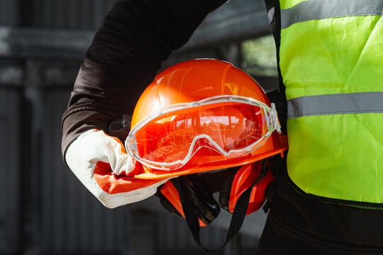 Man Worker holding orange safety helmet with protective goggles and earmuffs in reflective vest. Occupational health and safety compliance, personal protective equipment concept.