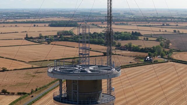 Close-up aerial view of the pinnacle and antenna array of a telecommunications transmitter mast over dry summer fields in the UK