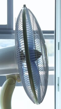 White electric pedestal fan spinning near a window during the day to help provide cool air circulation inside a room for comfort