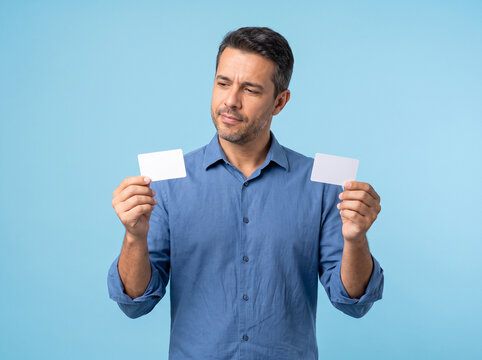 Thoughtful mature man holding two blank white cards making a choice between payment options on blue background