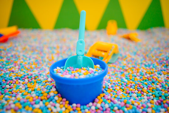 Various colorful plastic toys and shovels scattered in a sensory pit filled with multi colored foam beads at an indoor kids playground.