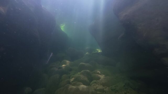 Low angle underwater view across smooth pebbles and large boulders on the bed of a deep river pool, with narrow sunbeams falling between dark rock walls in cool blue green water, calm and mysterious