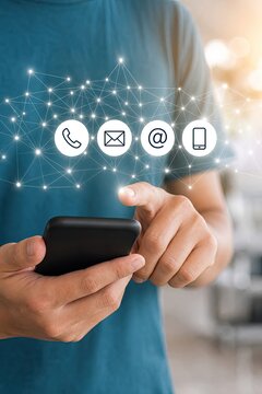 A close-up shot of a person's hands holding a black smartphone, with contact information icons for phone, email, and mobile floating above a blurred office background.