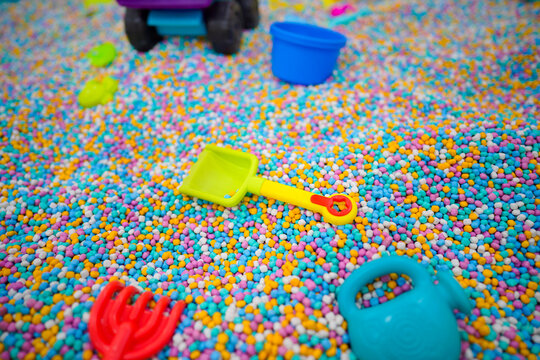 Various colorful plastic toys and shovels scattered in a sensory pit filled with multi colored foam beads at an indoor kids playground.