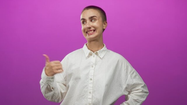 Woman with buzzcut gives a thumbs up showing thumb and teeth in a studio with magenta backdrop and white shirt; playful attitude.
