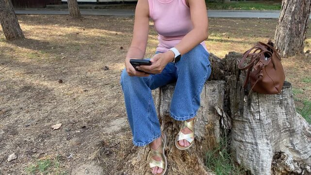 A woman sits on a tree stump while using her smartphone. Her brown handbag rests beside her in a quiet park. The peaceful moment brings a calm and relaxed feeling.