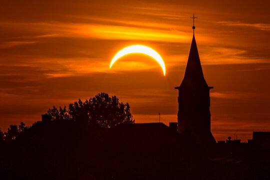 Partial solar eclipse above a church and silhouetted trees against a dramatic orange sunset sky, creating a striking celestial and atmospheric landscape.