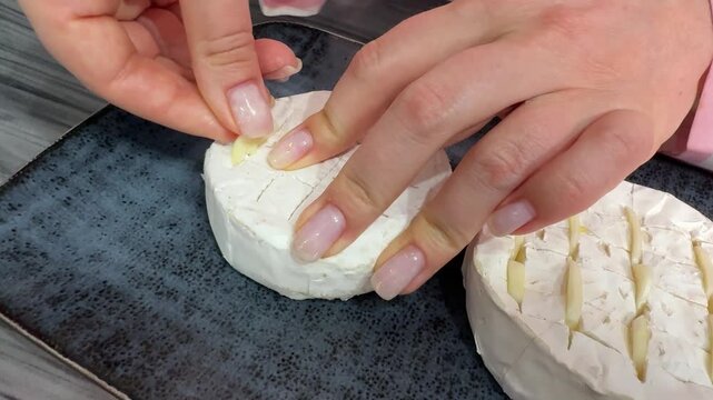 A female chef in the kitchen carefully stuffs a whole wheel of Brie cheese with fresh garlic slices, then bakes it in a hot oven until golden, soft, warm, and very gooey inside.