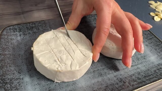 Female chef scores Brie cheese, inserts garlic slices, preps for baking until warm, melted, golden, and soft.