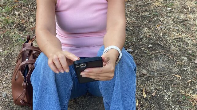 A woman sits outdoors while using her smartphone. Her brown handbag rests beside her on the dry ground in a park. The quiet break brings a calm and relaxed feeling.
