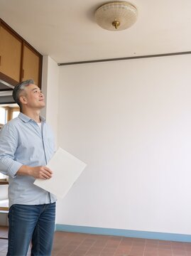 Asian man standing in empty room holding folder and looking up. Mature homeowner inspecting new house interior. Copy space