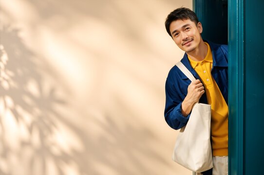 Smiling Asian man stepping out of a doorway holding a canvas tote bag. Modern lifestyle and eco friendly shopping concept. Copy space for text