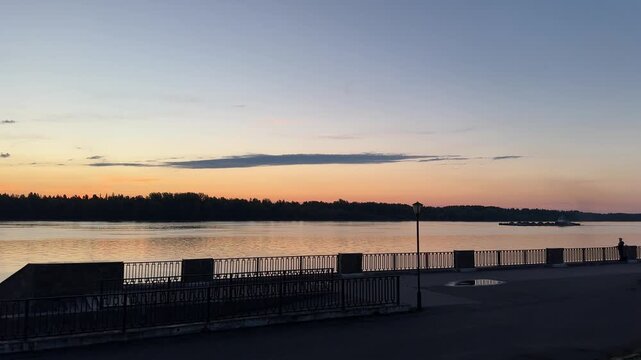 Dusk on the river promenade, a barge floats downstream, evening atmosphere, reflections on water, urban skyline, candid authentic life captured in a quiet moment.