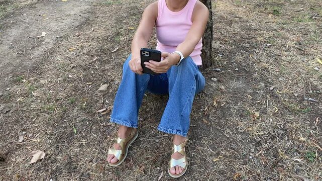 A relaxed woman sits on the ground leaning against a tree while holding her phone. She peacefully checks her screen in a quiet park setting. Her calm posture expresses contentment.