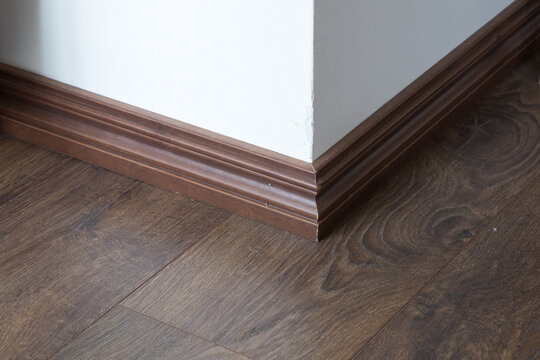 Classic brown wooden skirting board installed on a wall corner. The decorative baseboard finishes the edge between the dark laminate flooring and a white painted wall.
