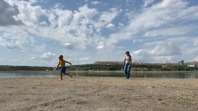 A young boy leans down to hit a shuttlecock while playing badminton with his mother. They enjoy their game by a peaceful lake under a cloudy sky. The fun moment brings warmth and joy.