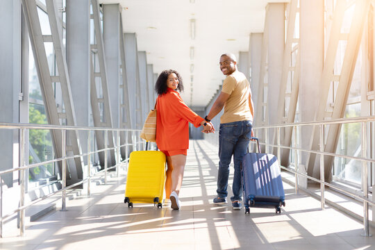 Smiling young Black couple looking back at camera while walking hand in hand through sunny airport terminal with rolling luggage on their upcoming holiday journey