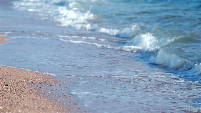 Close-up view of clear blue sea waves washing gently over a sandy and pebble beach shore with white foam