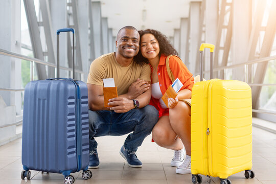 Happy couple crouching together between suitcases at airport, holding passports and boarding passes while smiling for the camera